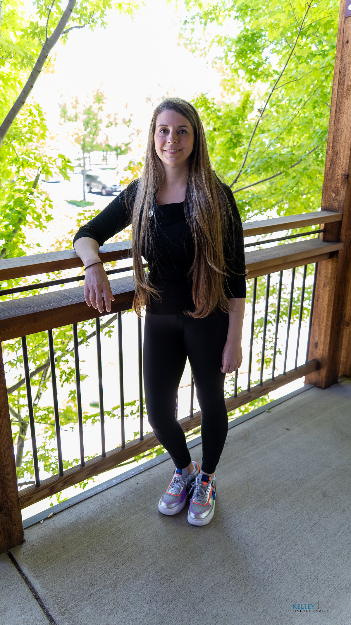 A woman with long brown hair stands on a porch, wearing a black outfit and colorful sneakers. Green trees and a wooden railing are in the background, creating a serene setting that reflects holistic approaches to dental care.