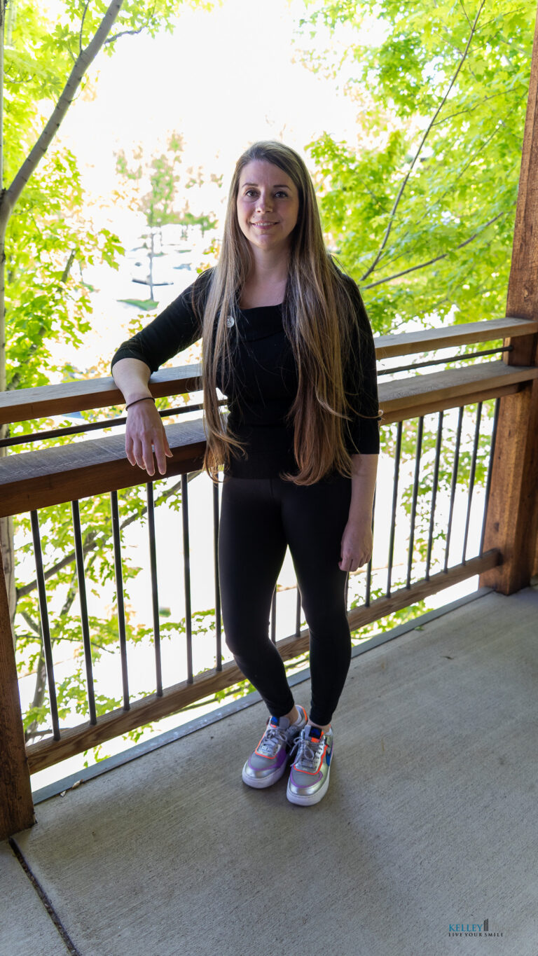 A woman with long hair stands on a balcony outside, leaning on a wooden railing in a black outfit and colorful sneakers. Green leaves frame the scene, evoking the freshness often found in holistic approaches to dental care.