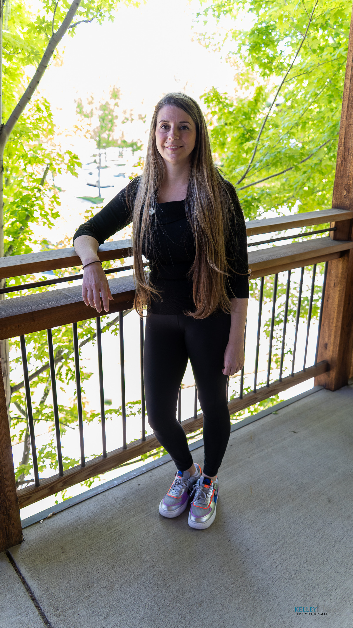 A woman with long hair stands on a balcony outside, leaning on a wooden railing in a black outfit and colorful sneakers. Green leaves frame the scene, evoking the freshness often found in holistic approaches to dental care.