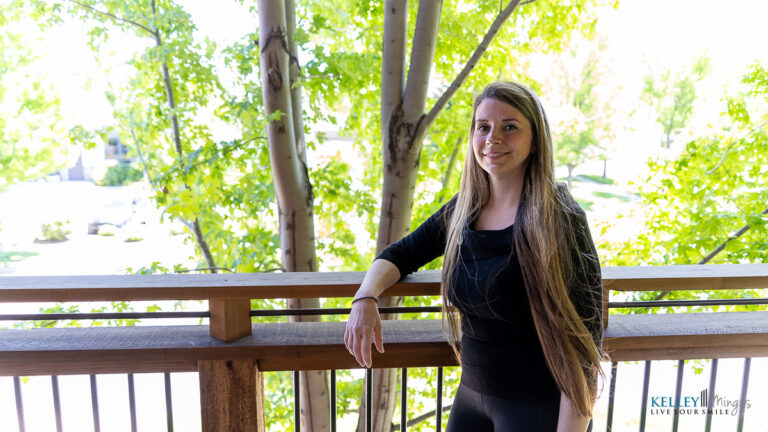 A woman with long hair stands on a wooden balcony outside, surrounded by green trees on a bright day, embracing holistic approaches to dental care and overall well-being.
