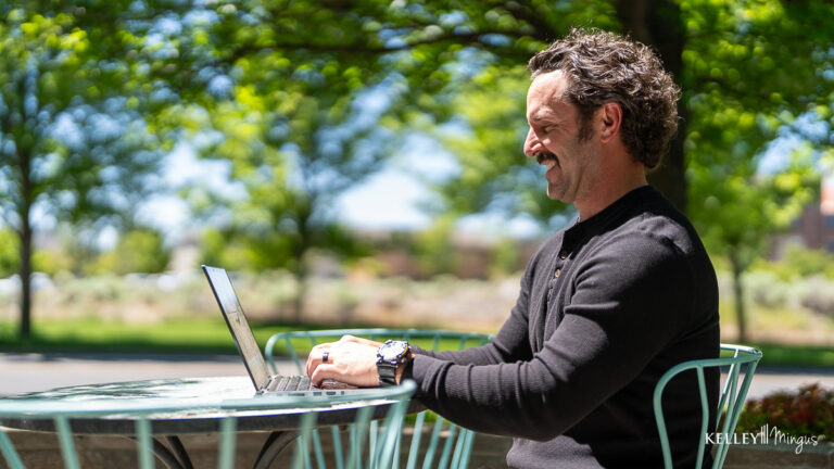 A man sits at an outdoor table, typing on a laptop, enjoying the tranquility of green trees and sunlight—perhaps researching holistic TMJ treatment options in this peaceful setting.
