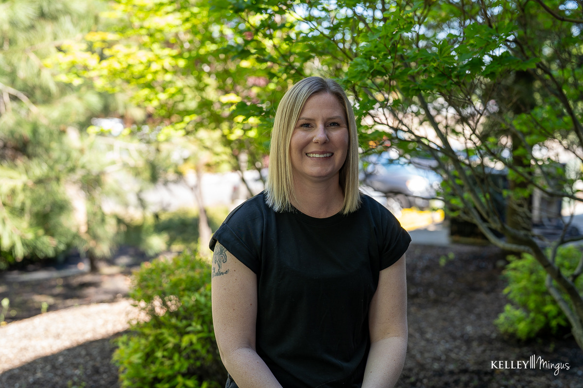 A person with straight blonde hair and a black t-shirt is sitting outside, smiling, surrounded by green foliage and sunlight, enjoying a bright day after visiting their family dentist.