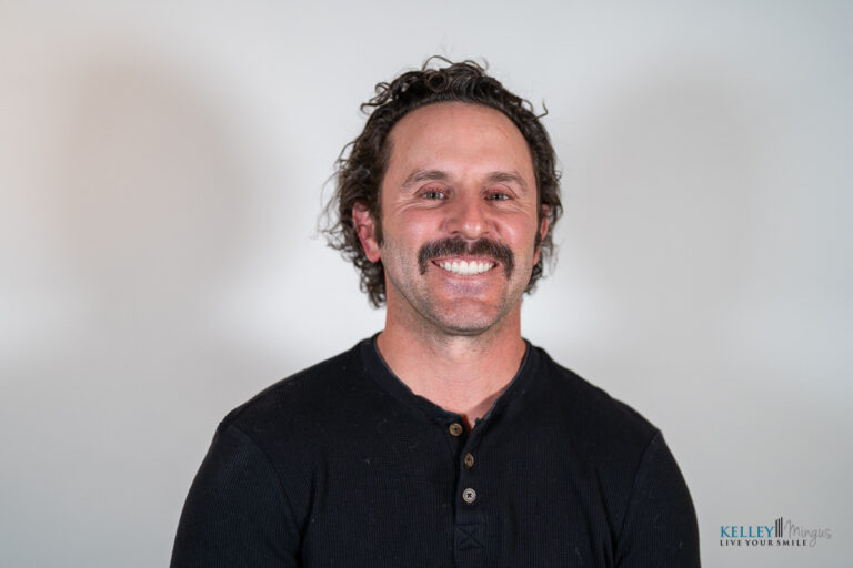 A man with curly hair and a mustache, wearing a black henley shirt, smiles at the camera against a plain light background, embodying confidence through effective TMJ pain management.
