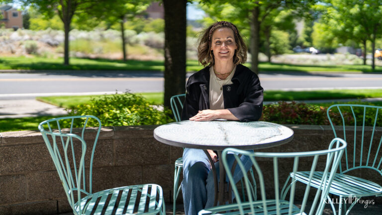 A woman with a bright smile, thanks to porcelain veneers for chipped teeth, sits alone at an outdoor metal table on a shaded patio, empty chairs around her and trees lining the street in the background.