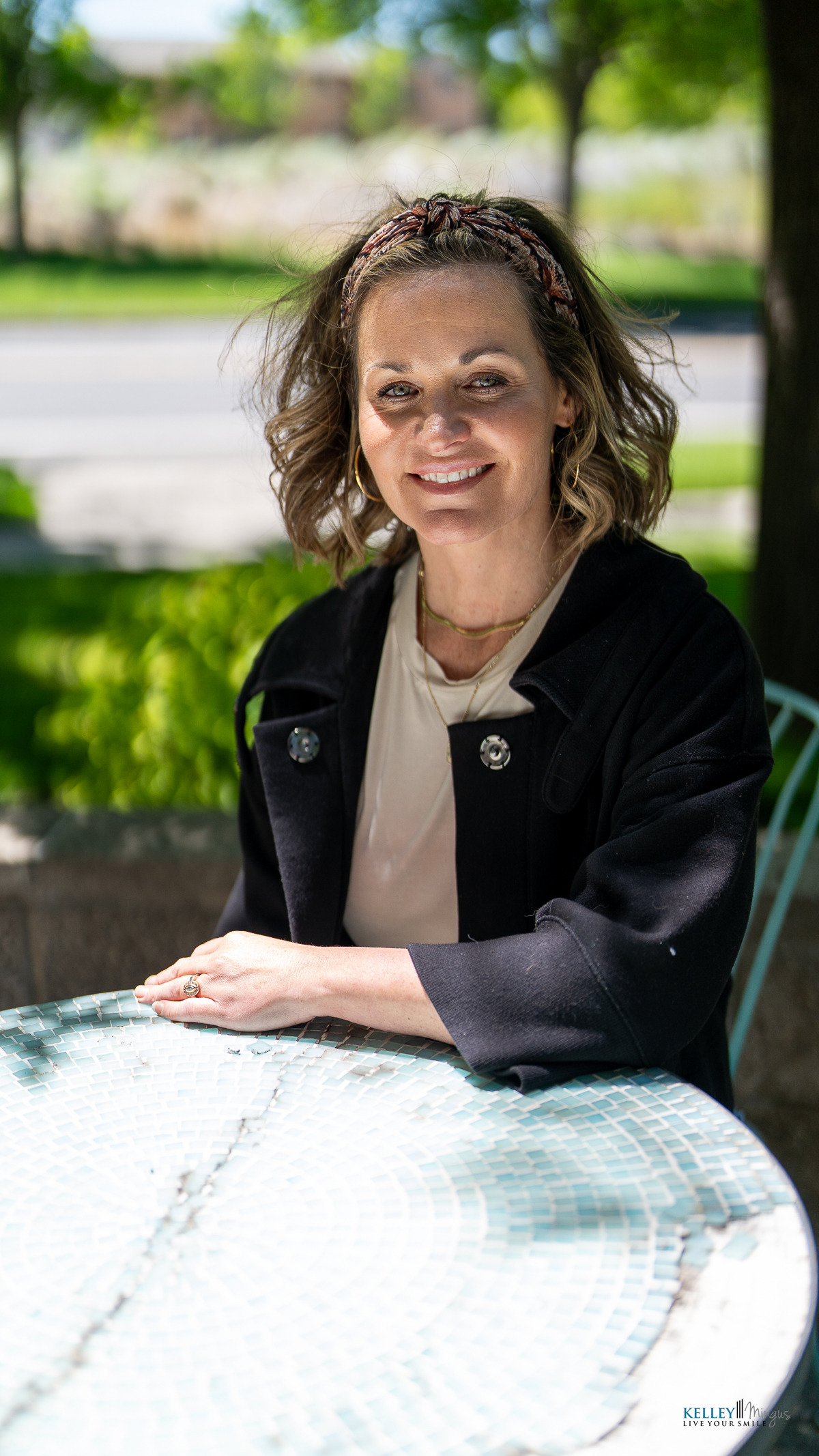 A woman with wavy hair and a headband sits at an outdoor table, smiling at the camera—a true picture of a healthier smile. Green trees and a road are visible in the background.