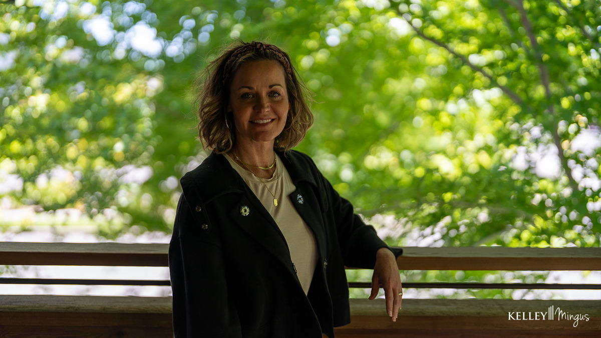 A woman stands on a wooden deck, smiling confidently at the camera, with green trees in the background. The image, watermarked "Kelley Mingus," highlights the boost from porcelain veneers for chipped teeth.