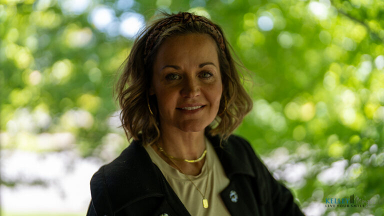 A woman with short, wavy hair wearing a black jacket and gold necklace stands outdoors with greenery in the background, reflecting confidence despite living with Binocular Vision Dysfunction (BVD).