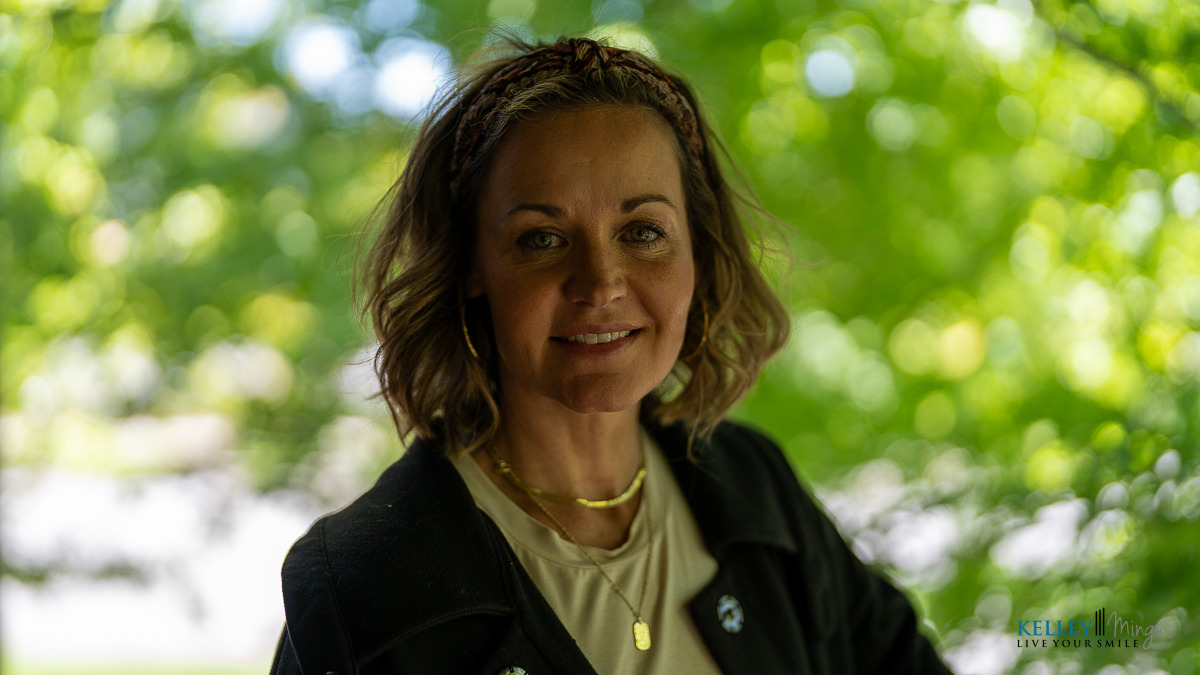 A woman with short, wavy hair wearing a black jacket and gold necklace stands outdoors with greenery in the background, reflecting confidence despite living with Binocular Vision Dysfunction (BVD).