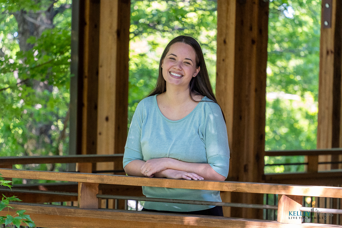 A woman with long brown hair, wearing a light blue top, stands and smiles while leaning on a wooden railing in an outdoor setting with green trees—an image often used in articles about living well with TMJ disorders or Binocular Vision Dysfunction (BVD).