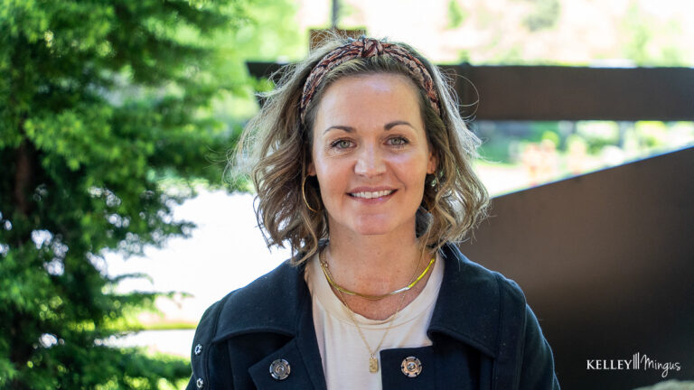 A woman with wavy, shoulder-length hair and a headband smiles outdoors, wearing a black jacket over a light shirt, with trees and a blurred structure in the background—a common scene among Bend residents embracing holistic dentistry.