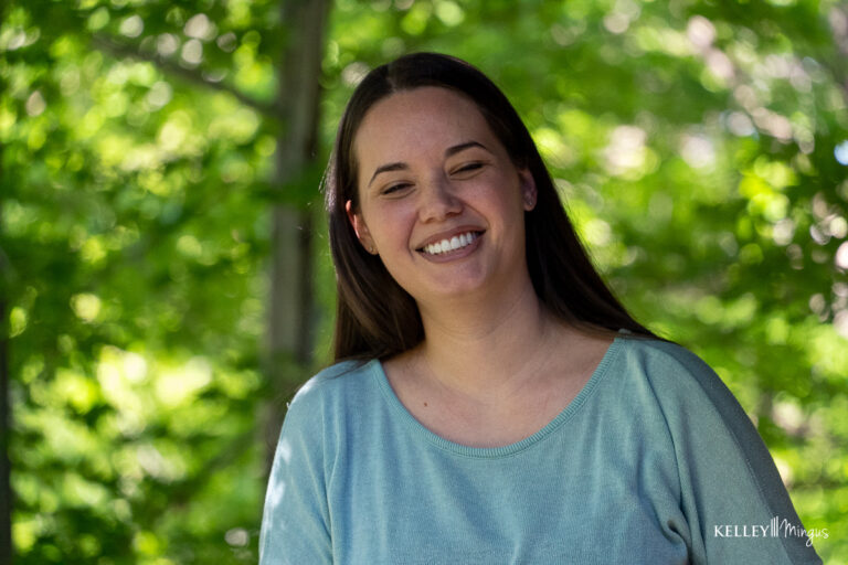 A woman with long brown hair, wearing a light blue top, smiles outdoors with green foliage blurred in the background, radiating positivity despite living with Binocular Vision Dysfunction (BVD).