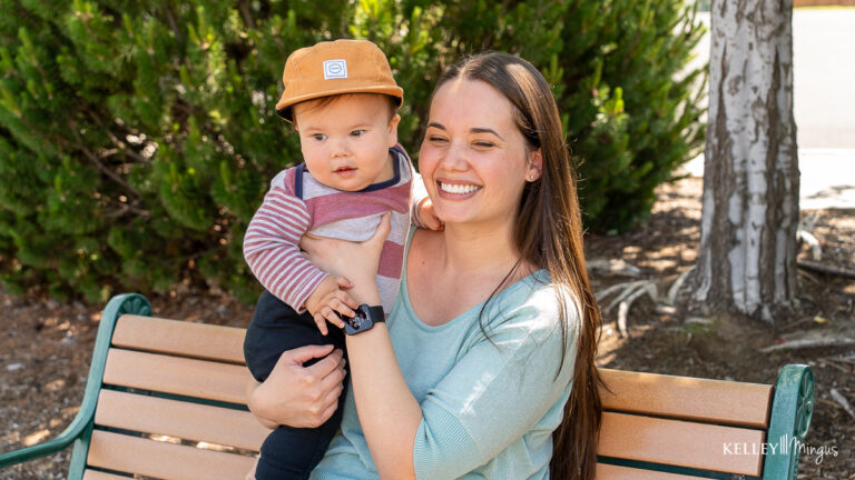 A woman sits on a bench outdoors, holding a baby in a brown hat and striped shirt. Both gaze slightly off camera, surrounded by greenery—a peaceful moment that highlights the importance of family time and dental care for seniors.