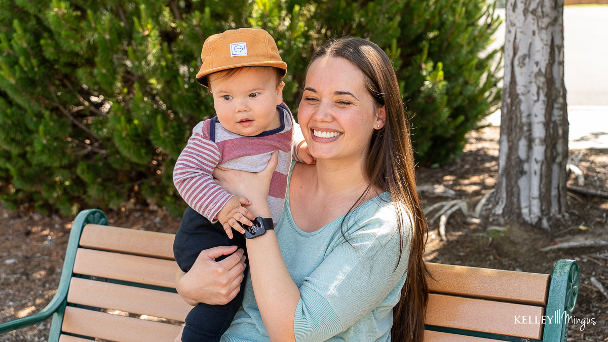 A woman sits on a bench outdoors, holding a baby in a brown hat and striped shirt. Both gaze slightly off camera, surrounded by greenery—a peaceful moment that highlights the importance of family time and dental care for seniors.
