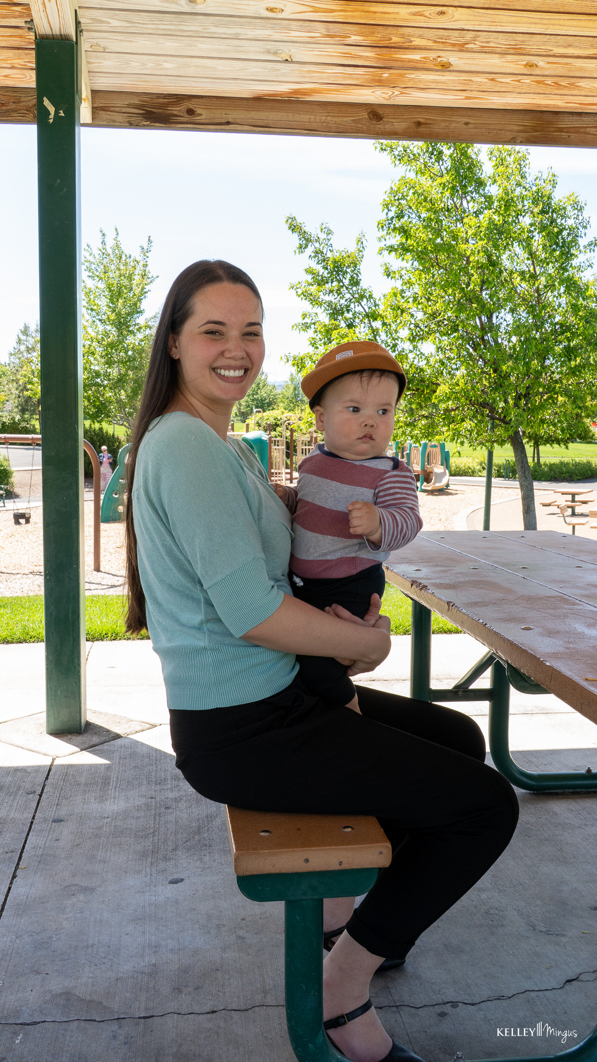 A woman sits on a picnic bench holding a baby in a yellow hat at an outdoor park with a playground in the background, enjoying fresh air and relaxation—important elements for TMJ pain management.