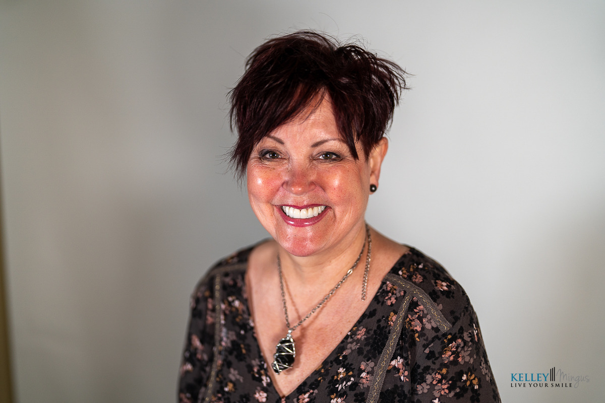A woman with short dark hair and a patterned top smiles at the camera against a plain light background, radiating confidence that comes from effective TMJ pain management.