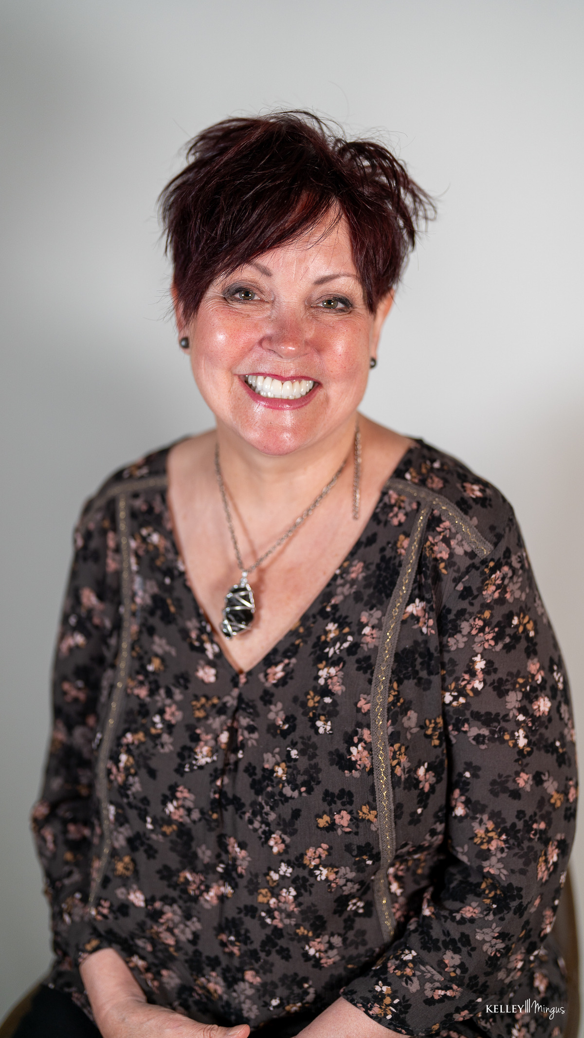 A woman with short dark hair, wearing a floral blouse and a necklace, smiles while sitting against a plain light background, embodying comfort and confidence gained from understanding sleep apnea symptoms.
