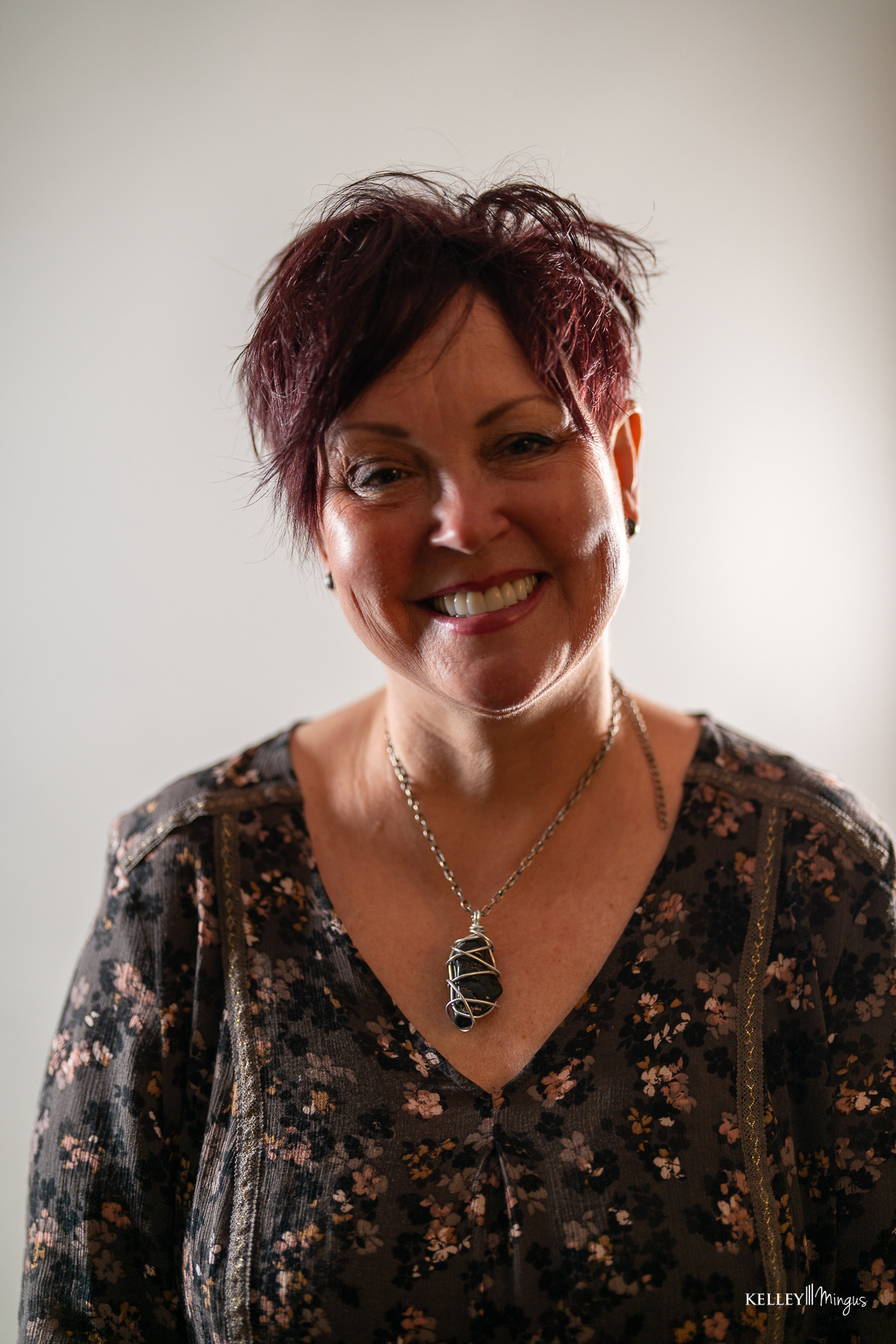 A woman with short, dark red hair and a patterned blouse flashes a custom smile design at the camera, wearing a large wire-wrapped stone necklace.