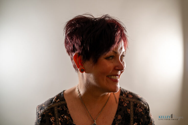 A woman with short, dark red hair smiles and looks to her left, showcasing a custom smile design. She wears a floral top, chain necklace, and stud earrings. The background is softly lit and neutral.