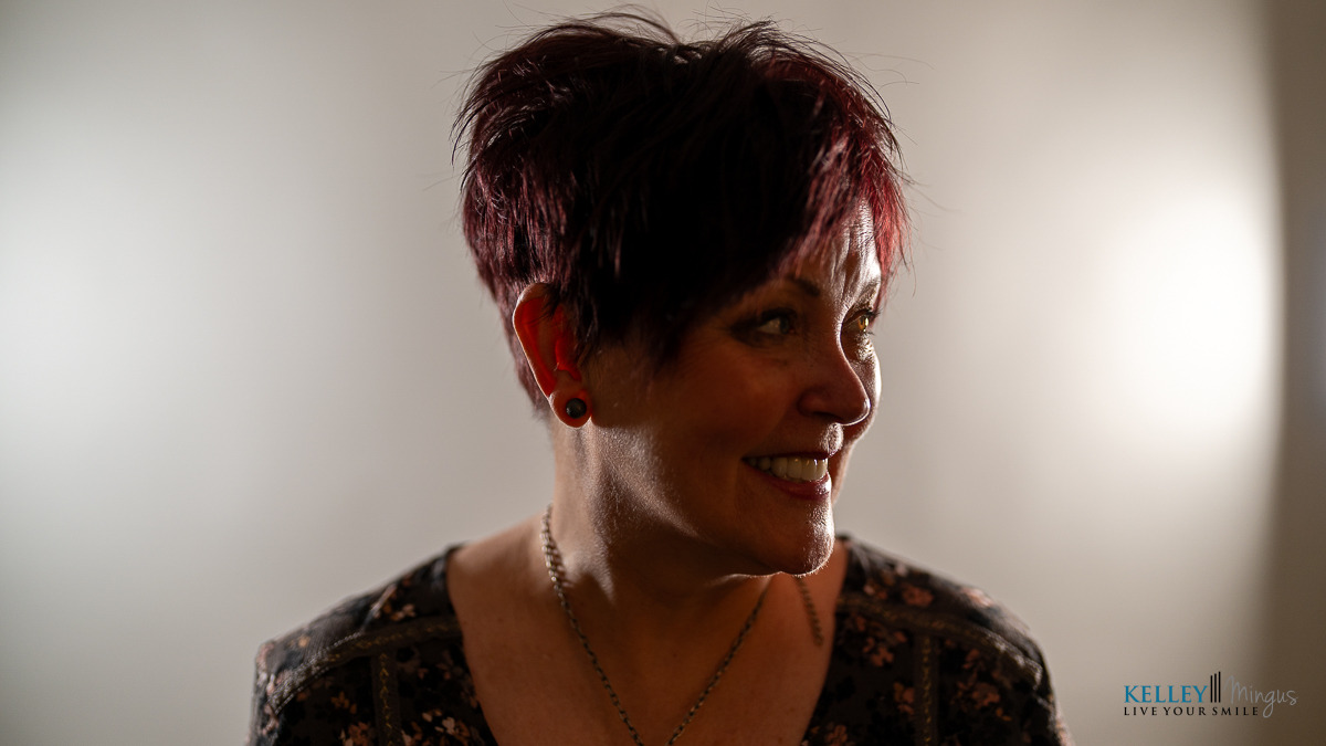 A woman with short, dark red hair smiles and looks to her left, standing in front of a neutral backdrop with soft lighting, her confidence shining after getting porcelain veneers for chipped teeth.