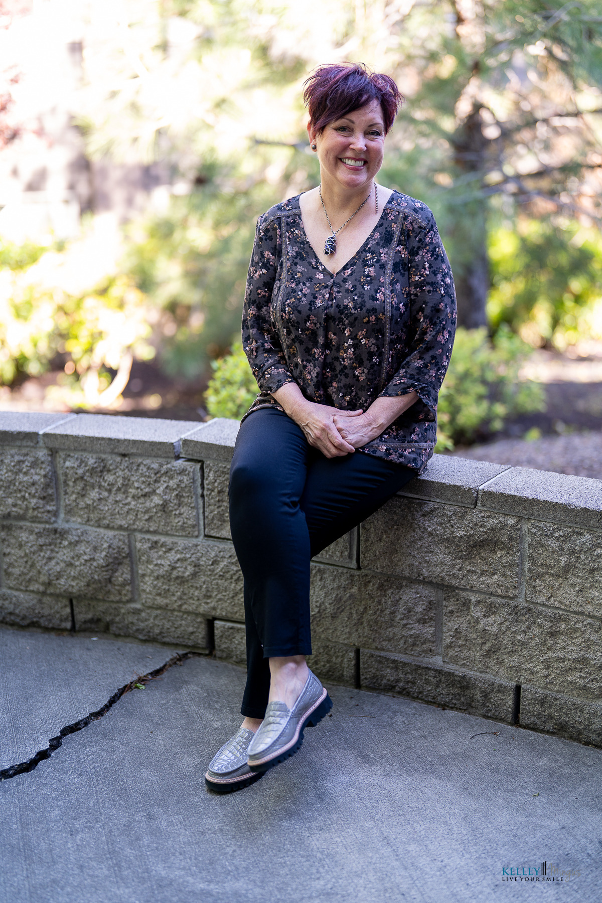 A woman with short hair sits on a low stone wall outdoors, showing off her custom smile design while smiling at the camera. She wears a patterned blouse, dark pants, and loafers, with greenery and trees in the background.