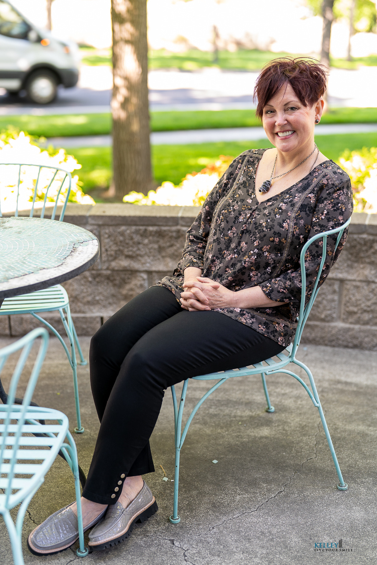 A woman with short hair, wearing a floral blouse and black pants, sits and smiles with a custom smile design on a light blue metal chair at an outdoor patio table.