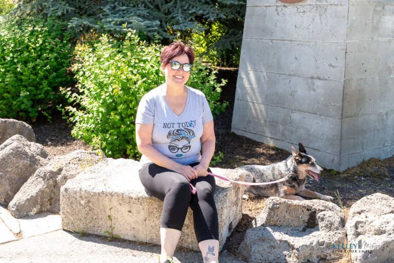 A woman in sunglasses sits on a stone bench outdoors with a small dog on a leash. She is wearing a "NOT TODAY" t-shirt and flashes a custom smile design while smiling at the camera.