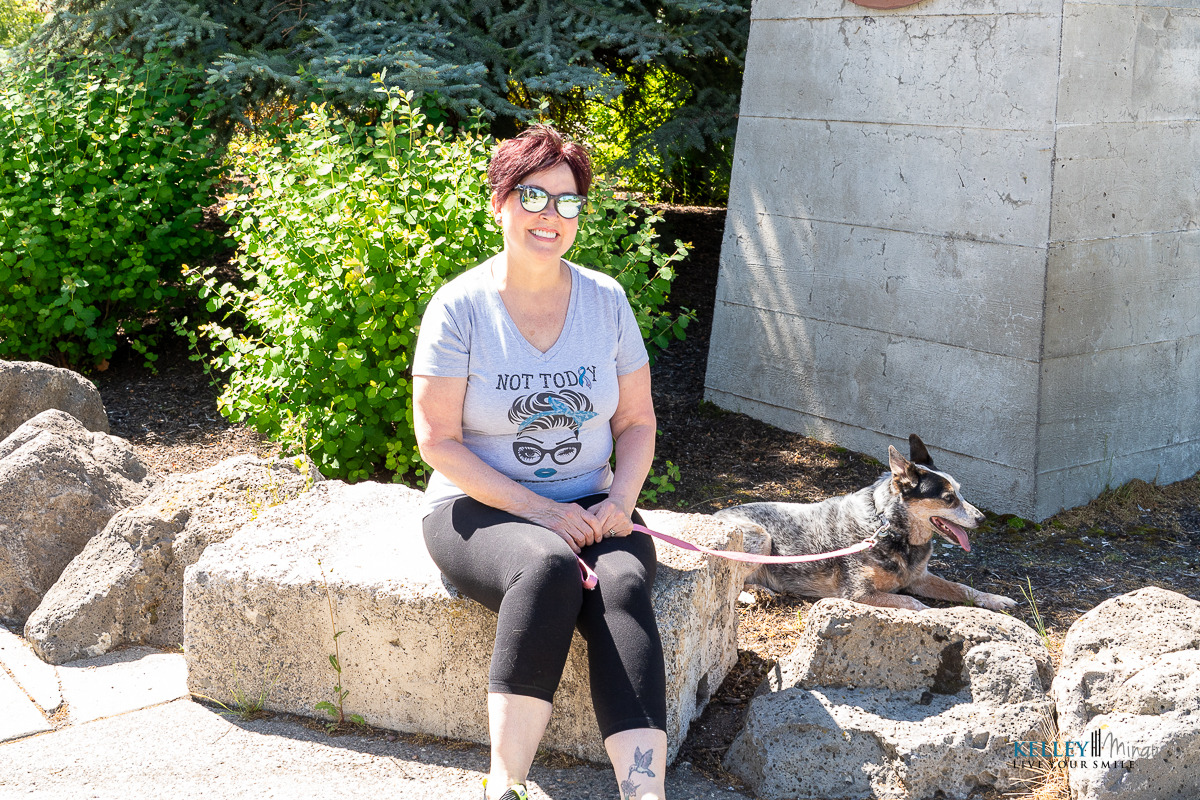 A woman in sunglasses sits on a stone bench outdoors with a small dog on a leash. She is wearing a "NOT TODAY" t-shirt and flashes a custom smile design while smiling at the camera.