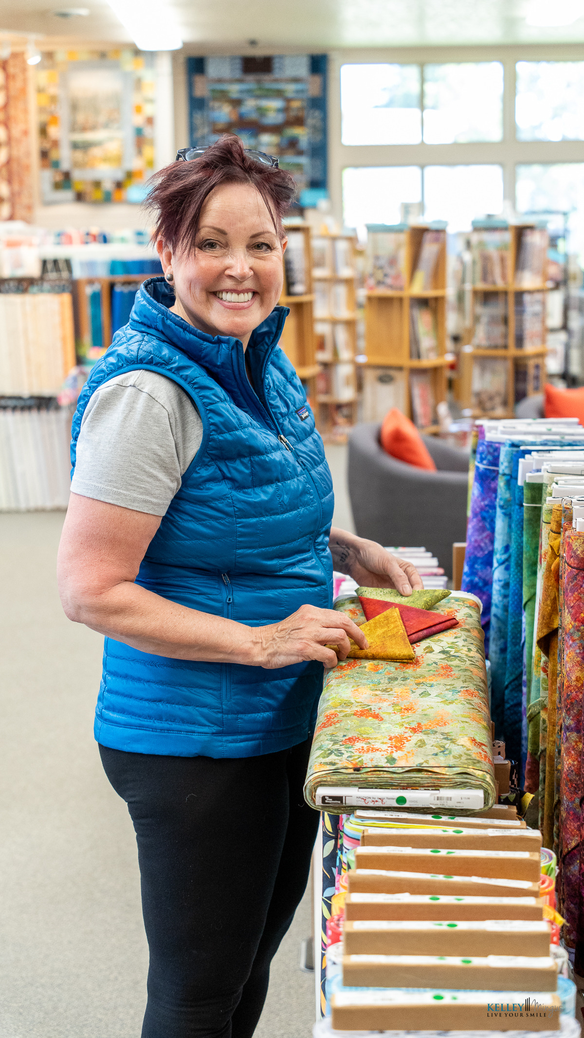 Woman in a blue vest, showcasing a custom smile design, smiles while selecting colorful fabric bolts at a fabric store.