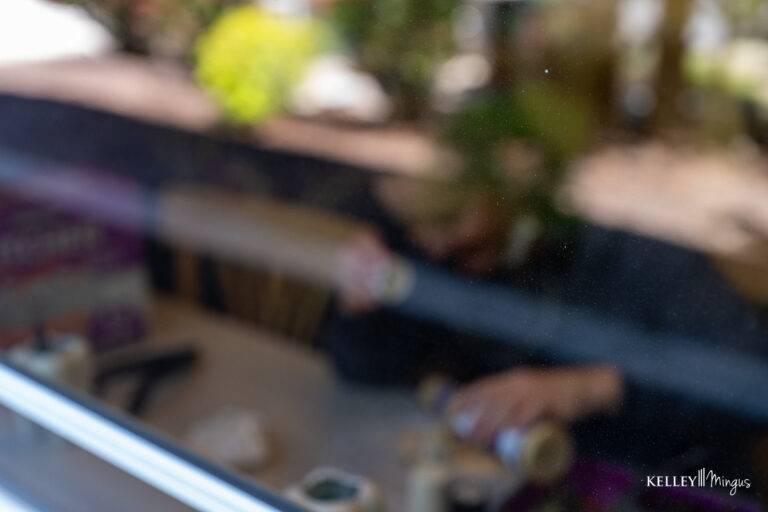 A blurry view through a window shows a person sitting at a table with various items, including cans and materials related to TMJ pain management, partially visible in the foreground.