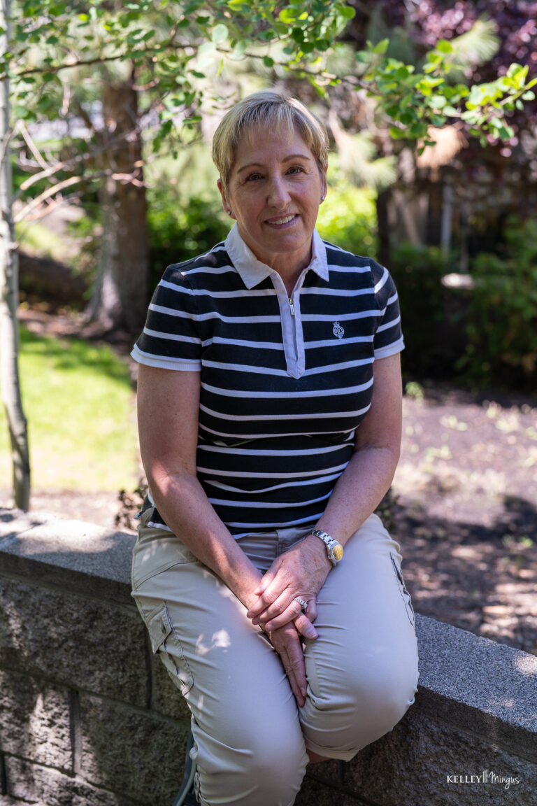 A woman with short blond hair sits on a stone wall outdoors, wearing a black-and-white striped polo shirt and beige pants, her radiant expression suggesting the confidence of a custom smile design, with lush greenery in the background.