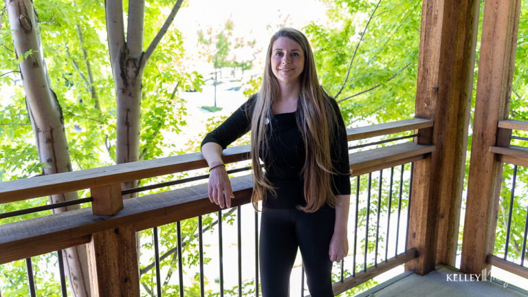 A woman with long brown hair stands on a wooden balcony surrounded by green trees, wearing a black outfit. The name "Kelley," who is also a trusted family dentist, appears in the lower right corner.
