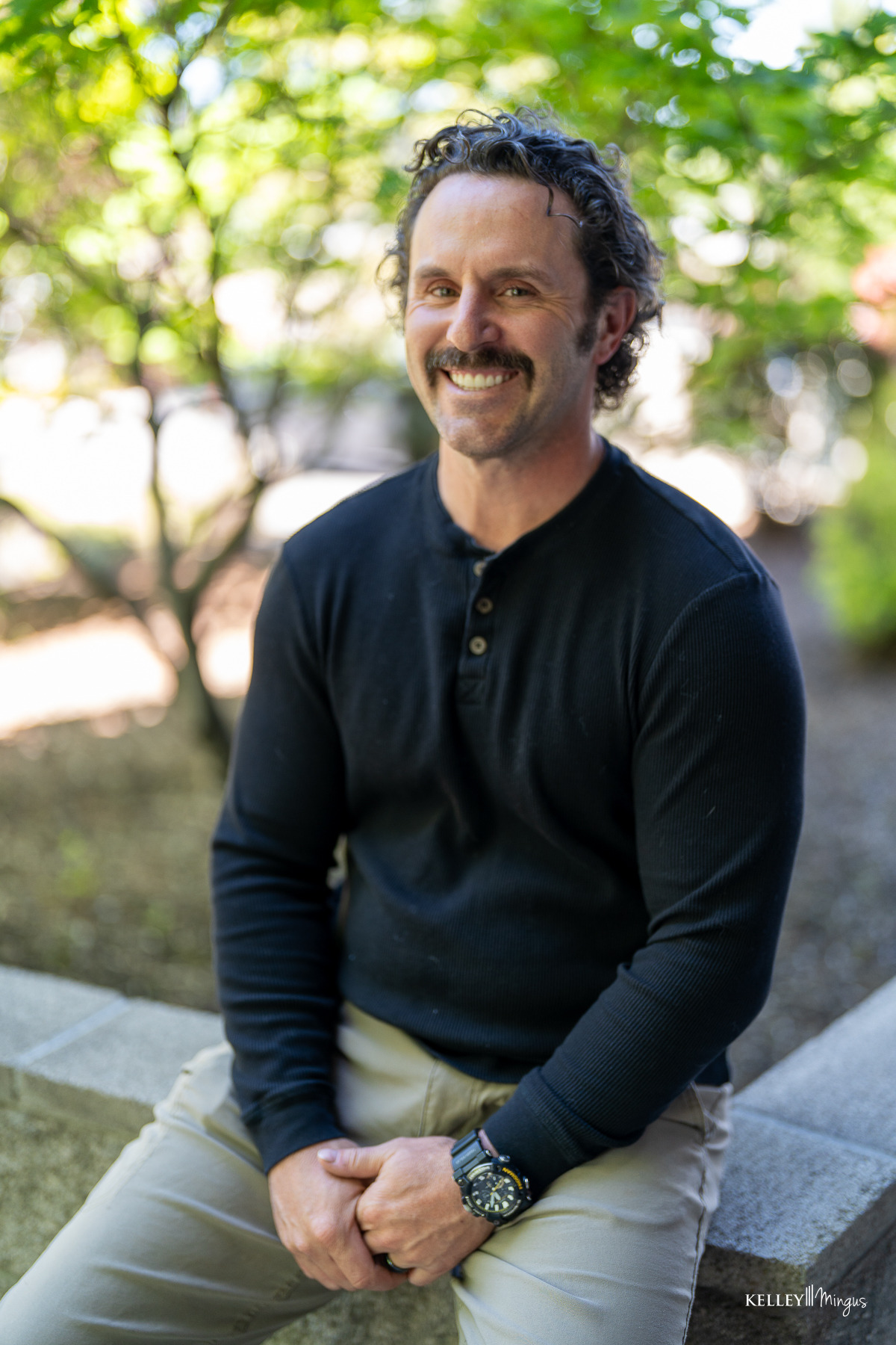 A man with curly hair and a mustache, wearing a black long-sleeve shirt and khaki pants, sits outdoors on a stone ledge, smiling at the camera as he discusses TMJ pain management.