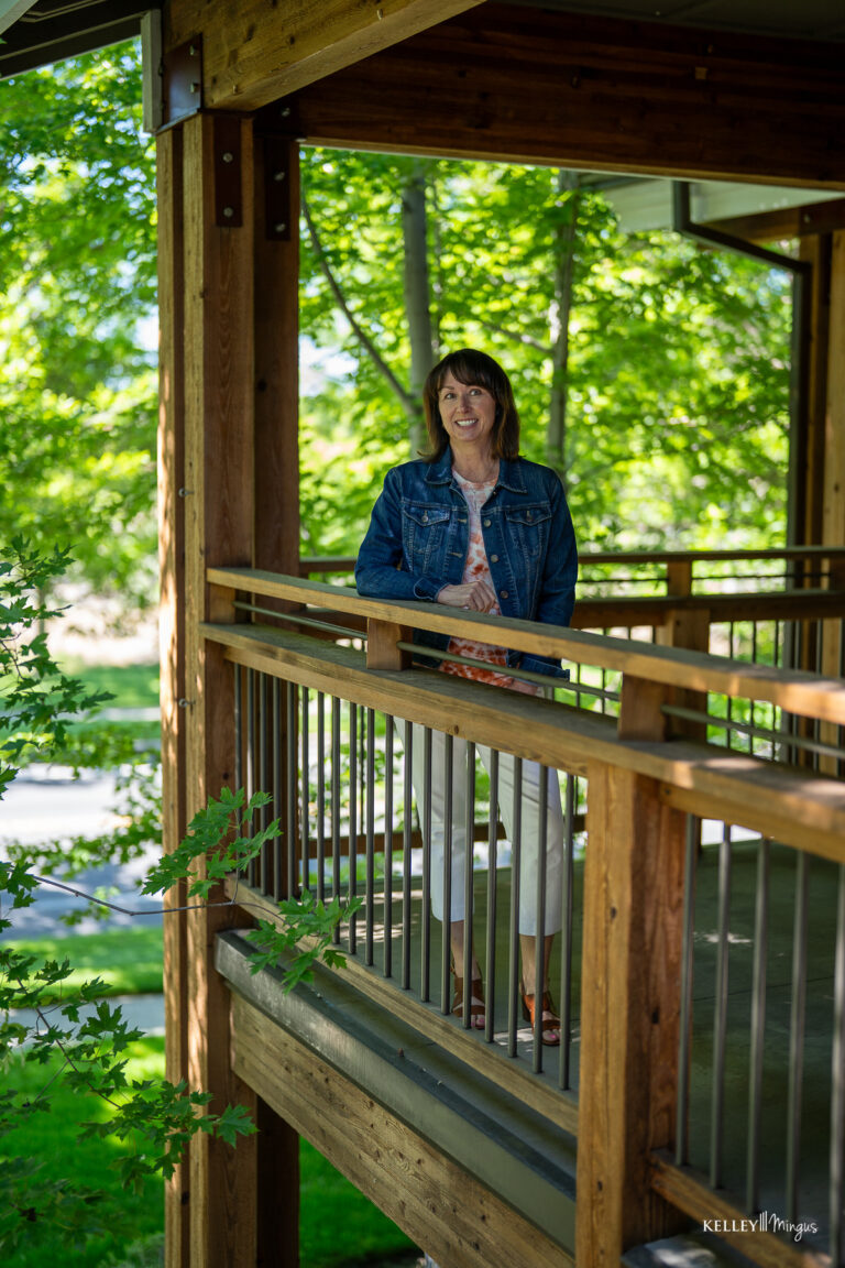 A woman stands and smiles on a wooden balcony surrounded by green trees on a sunny day, enjoying relief after TMJ treatment for migraines.