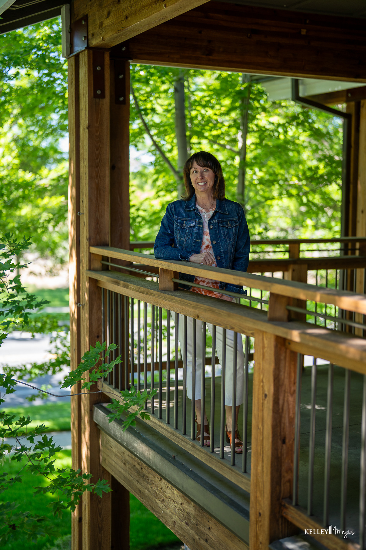 A woman stands and smiles on a wooden balcony surrounded by green trees on a sunny day, enjoying relief after TMJ treatment for migraines.