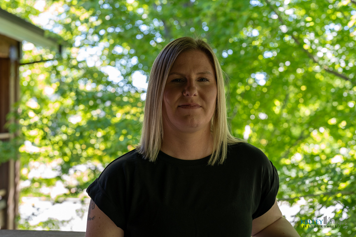 A woman with straight blonde hair and a black t-shirt stands outdoors in front of green leafy trees, with sunlight filtering through the branches, embodying the calm confidence that holistic approaches to dental care can inspire.