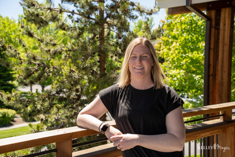 A woman with blonde hair in a black t-shirt stands outside, leaning on a wooden railing with trees and greenery in the background, enjoying the fresh air after her appointment for dental care for seniors in Bend.