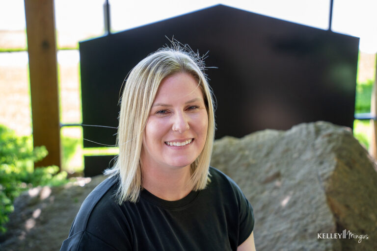 A woman with straight blonde hair and a black shirt smiles outdoors, seated in front of large rocks and a dark geometric structure, enjoying the fresh air after her appointment for dental care for seniors in Bend.