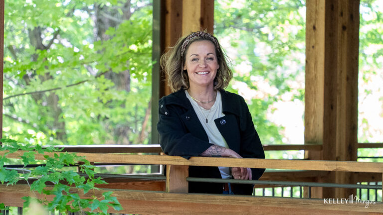 A woman stands and smiles at a wooden railing in an outdoor gazebo, surrounded by green trees and natural light—radiating happiness, much like the confidence brought by quality dental care for seniors in Bend.