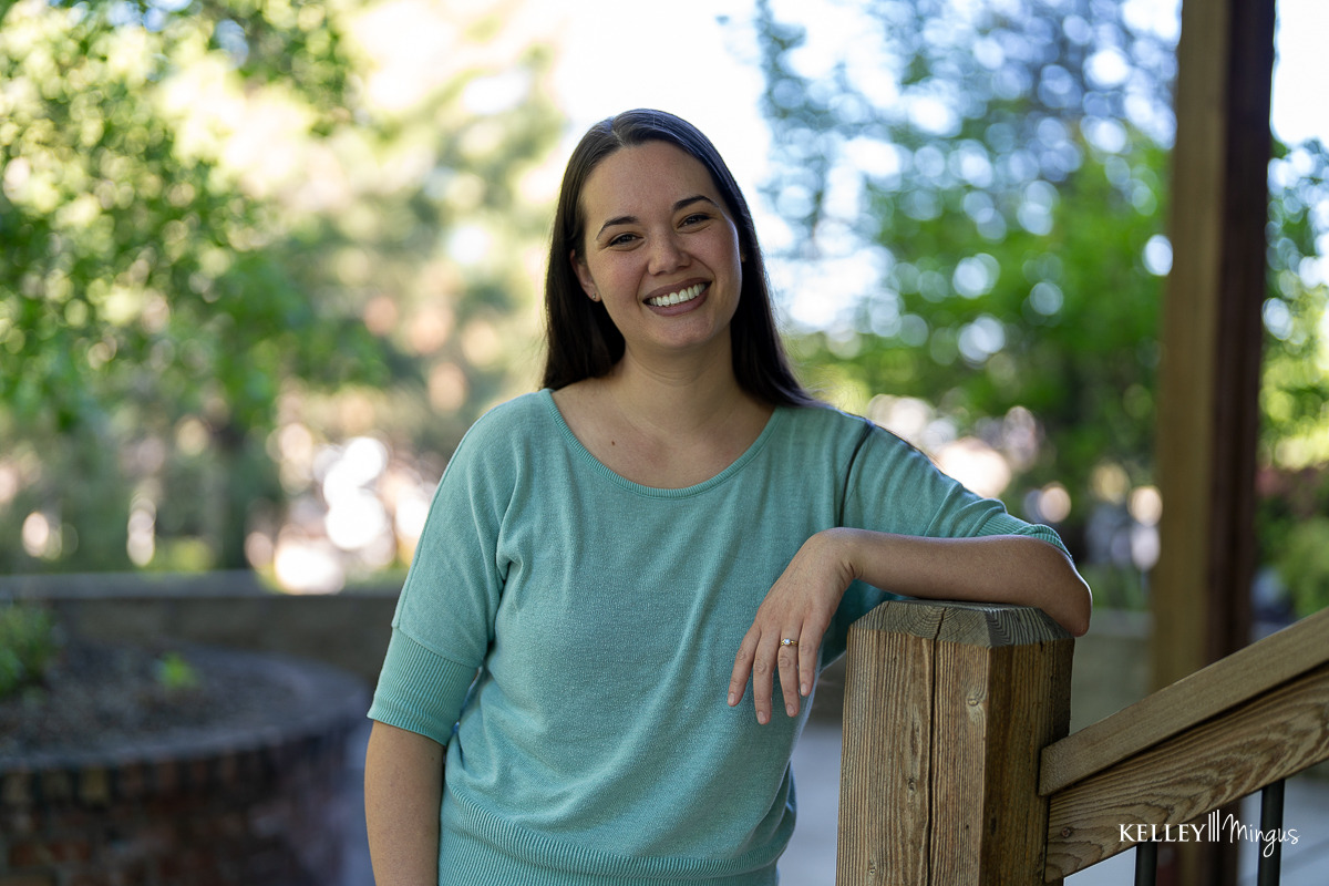 A woman with long brown hair, wearing a light blue top, smiles while leaning on a wooden railing outdoors with greenery in the background, showcasing her radiant smile after a Porcelain Veneers Consultation.