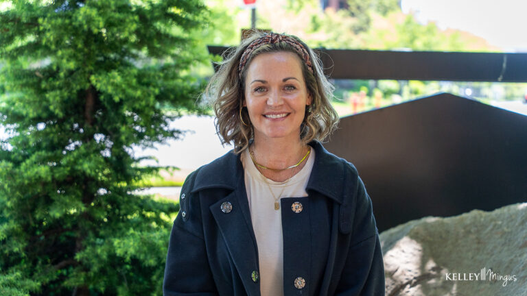 A woman with wavy hair, wearing a headband, light shirt, and dark jacket, smiles confidently at the camera while standing outdoors near greenery and rocks—her bright smile enhanced by metal-free restorations.