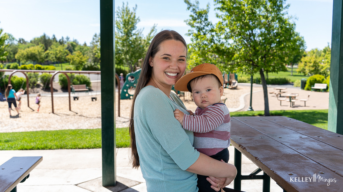Woman holding a baby under a park shelter, with playground equipment and people in the background on a sunny day, discussing metal-free dental restoration options.