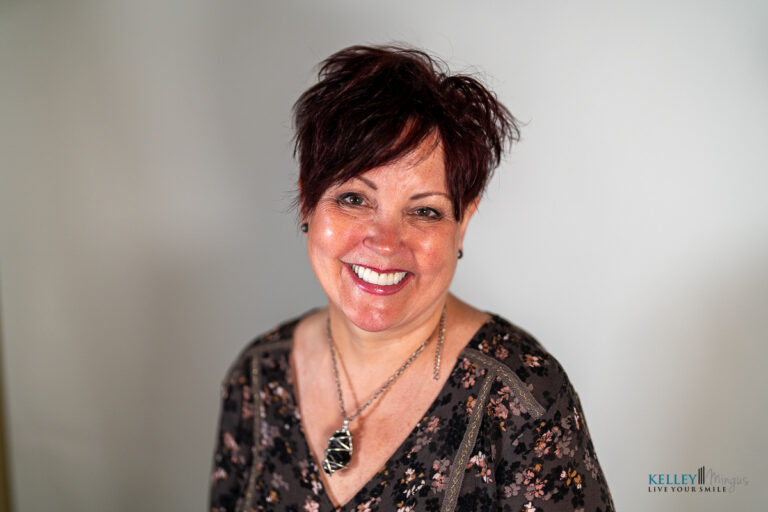 A woman with short dark hair and a patterned top smiles at the camera against a plain background, showcasing the confidence achieved through cosmetic dentistry in Bend, Oregon.
