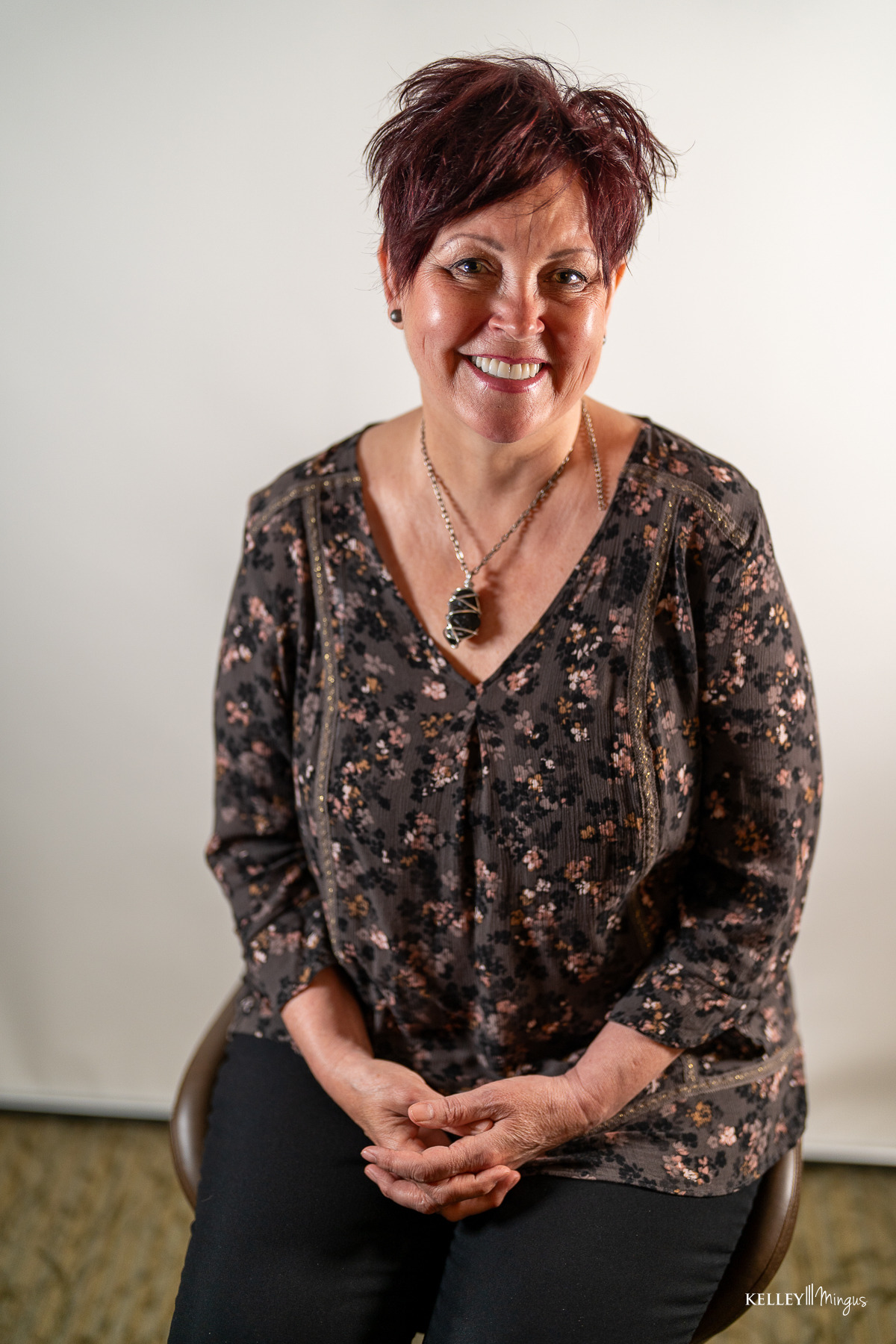 A woman with short dark hair wearing a floral blouse and pendant necklace smiles while sitting on a stool, embodying confidence and comfort—reflecting the benefits of a holistic approach to TMJ relief—against a plain background.