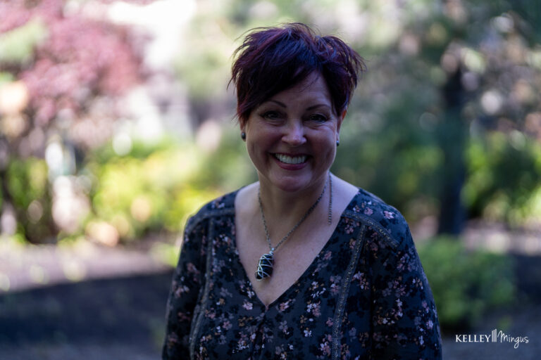 A woman with short dark hair, wearing a floral top and leaf-shaped necklace, smiles outdoors with greenery in the blurred background, enjoying life after exploring sleep apnea treatment options in Bend.