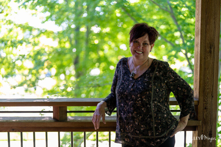 A woman stands on a wooden porch, smiling at the camera, with green trees in the background—enjoying life after exploring sleep apnea treatment options in Bend.