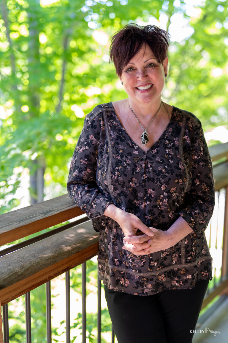 A woman with short dark hair stands and smiles on a wooden deck, wearing a floral blouse and necklace, with green trees in the background—enjoying life thanks to effective sleep apnea treatment options in Bend.