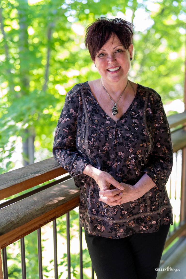 A woman with short dark hair stands outside on a wooden balcony, smiling confidently at the camera after a Full Mouth Rehabilitation, wearing a patterned blouse and necklace. Green trees are visible in the background.