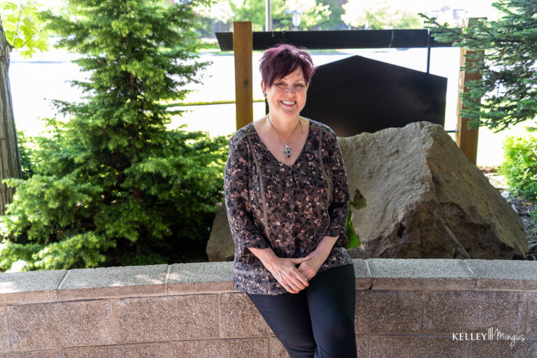 A woman with short, reddish hair smiles while sitting on a low brick wall outdoors, surrounded by greenery and large rocks—a peaceful moment that reflects her journey with holistic sleep apnea treatment.