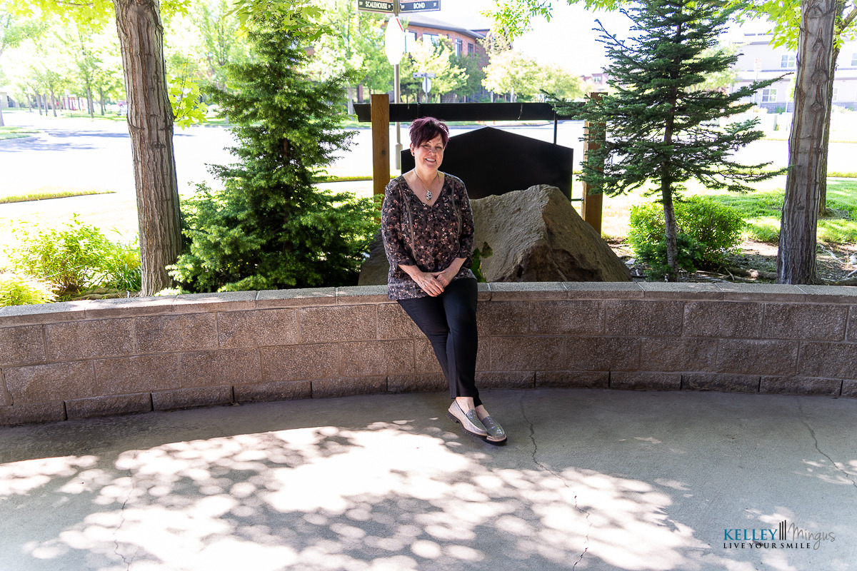 A woman sits on a curved stone bench in a shaded outdoor area, surrounded by trees, plants, and a large rock—inviting you to wonder: What alternatives might this peaceful setting inspire?.