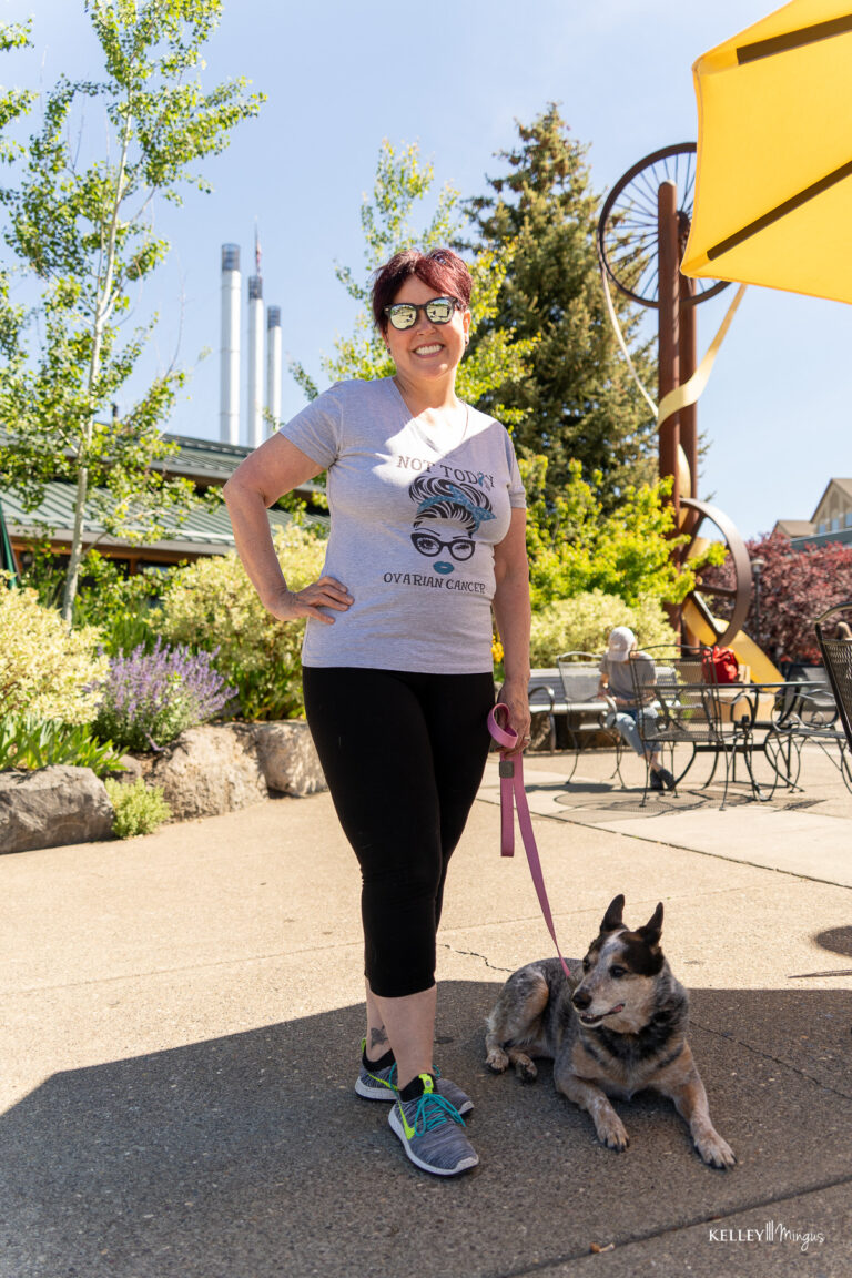A woman wearing sunglasses and a graphic t-shirt stands outside on a sunny day, holding a dog on a leash. Industrial chimneys and outdoor seating are visible in the background, reflecting the city’s vibe as fresh as the latest cosmetic dentistry trends.